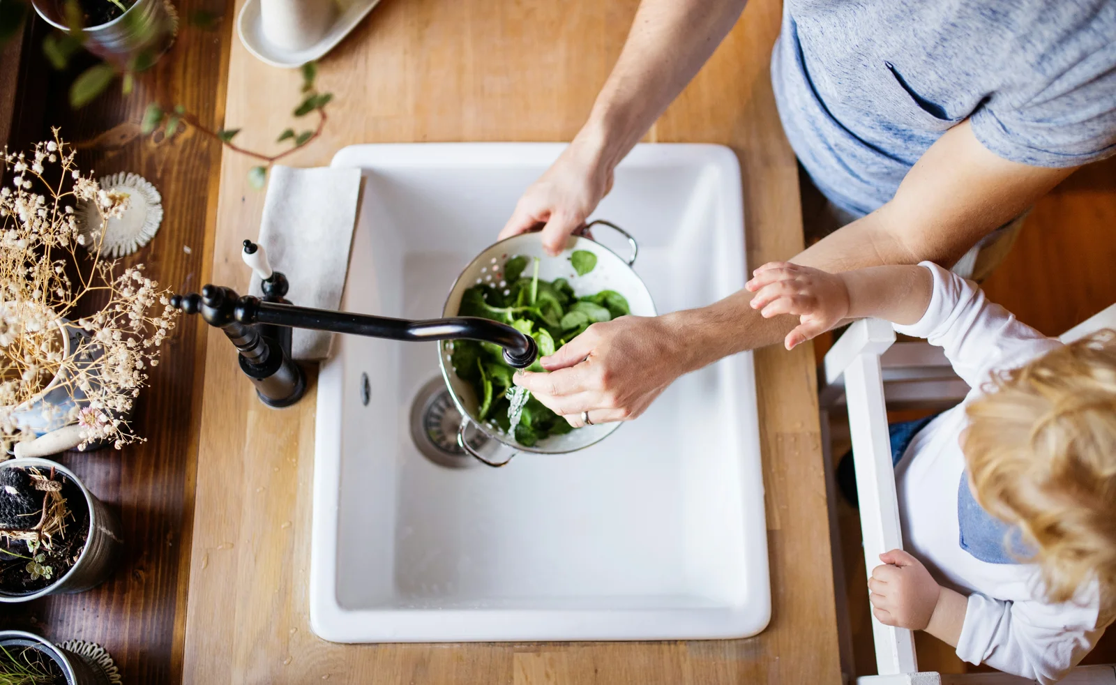 Parent and toddler rinsing greens at the kitchen sink
